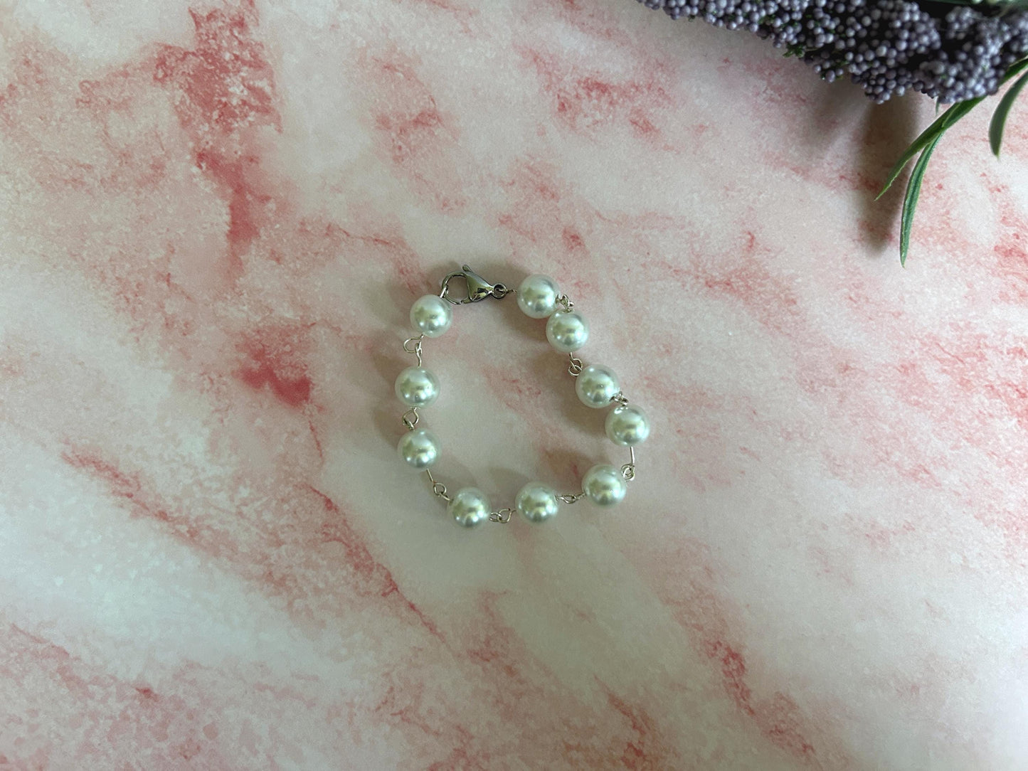 Pearl link bracelet clasped on a pink marble background with flowers peaking in the background.