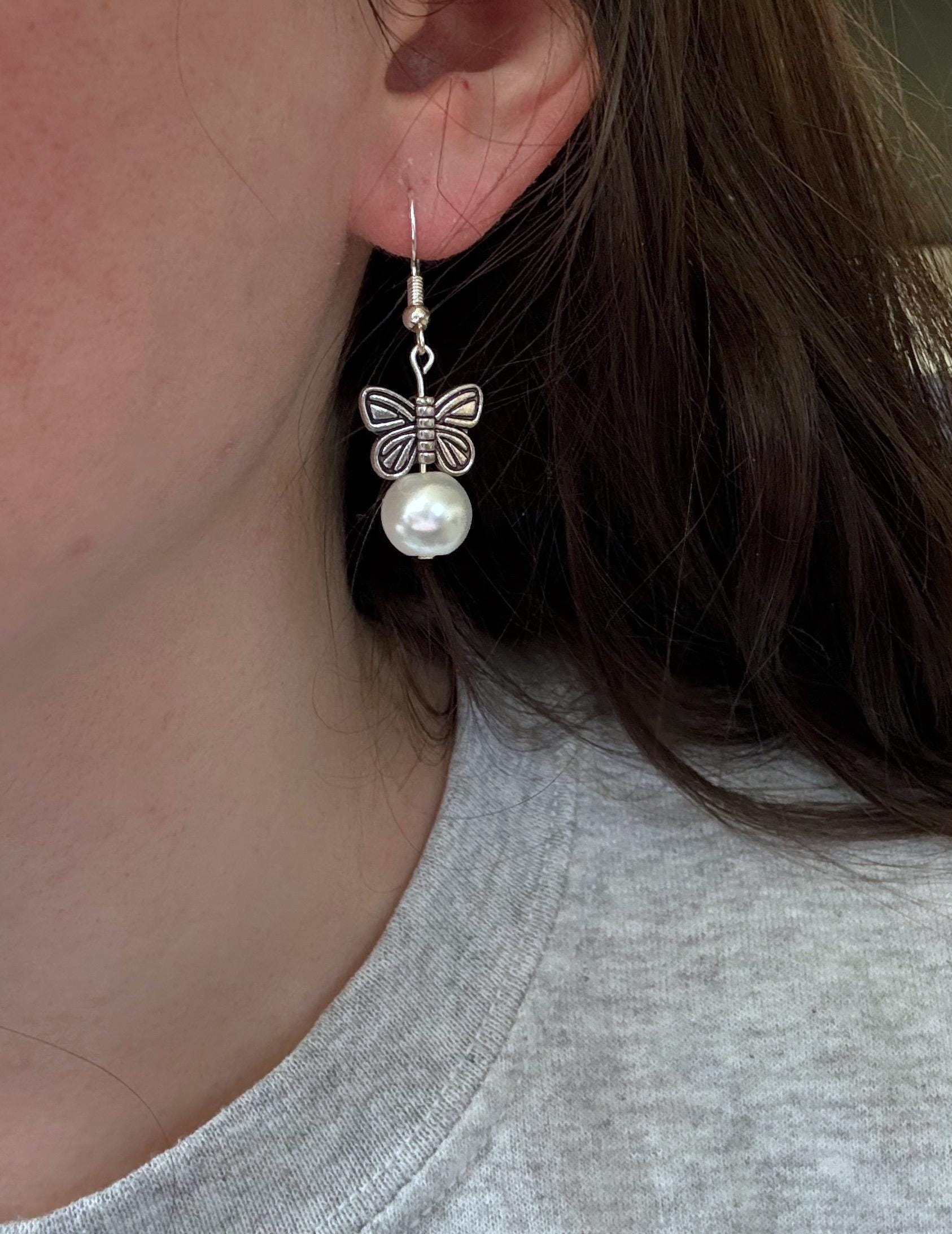 A pair of silver earrings butterfly earrings in an ear. The earrings feature metal butterflies and a pearl under the butterfly.