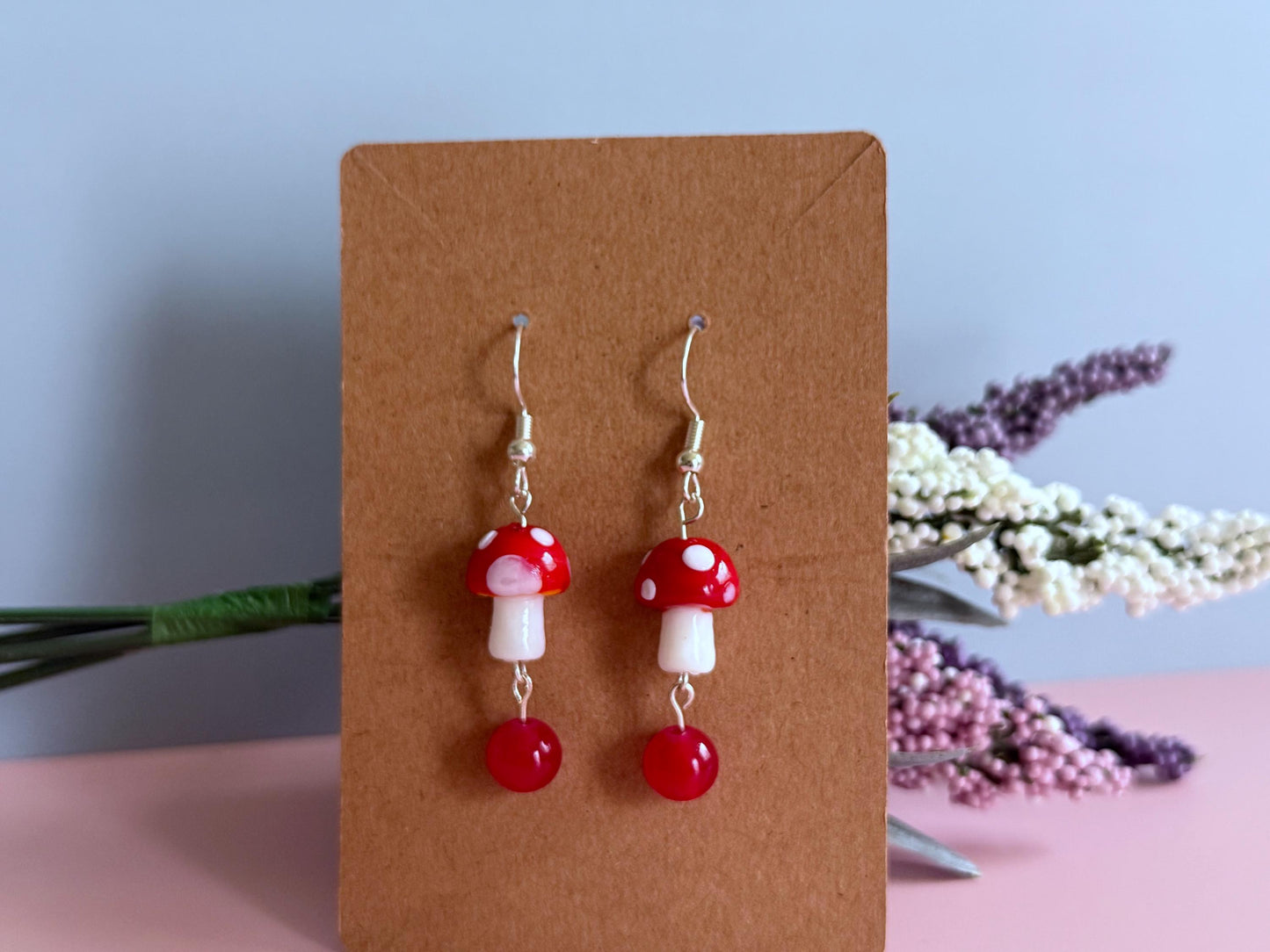 Red and white mushroom dangling earrings in silver with a red ball on the end on an earring card propped up on flowers.