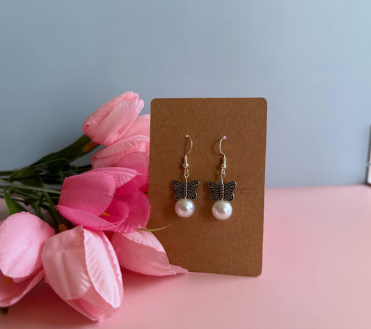 A pair of silver earrings butterfly earrings on an earring card with tulips beside it. The earrings feature metal butterflies and a pearl under the butterfly.