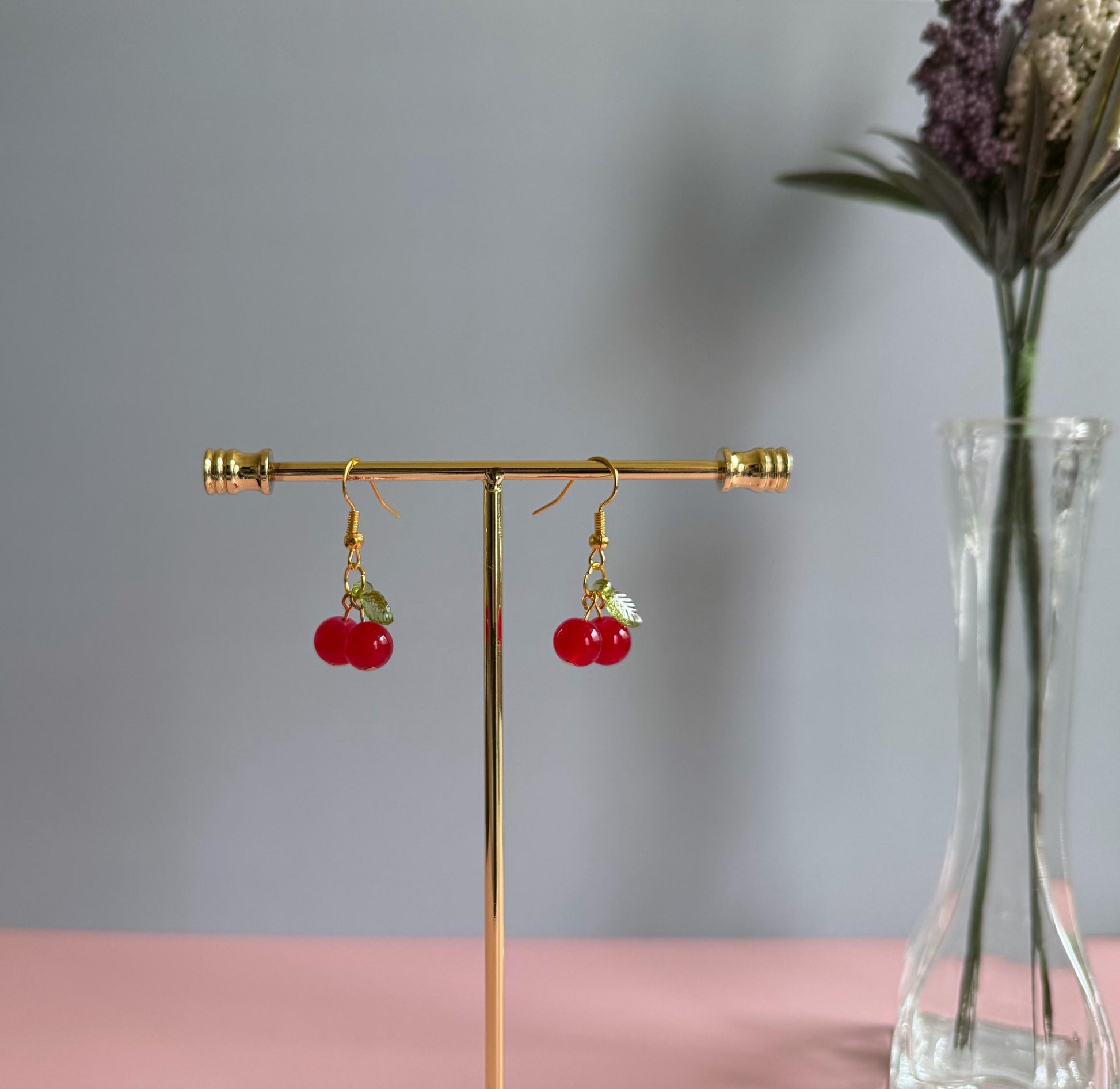 A pair of silver cherry earrings hanging from a stand with flowers in the background. The earrings feature shiny red coated glass beads and acrylic green leaf beads.