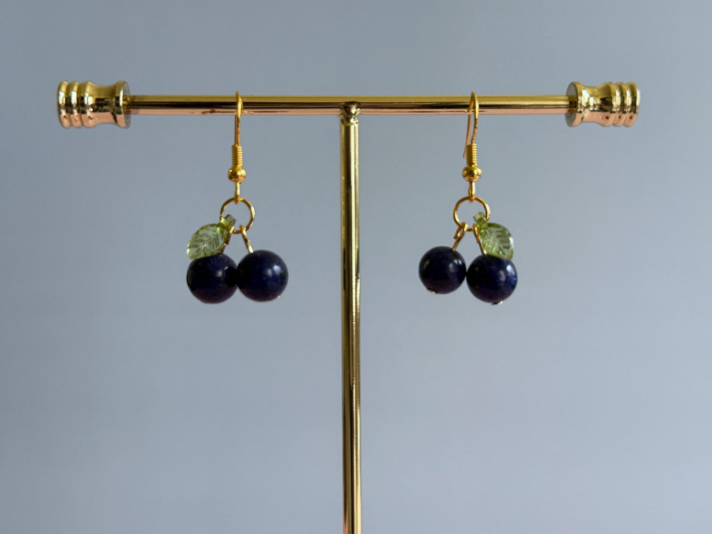 A pair of gold blueberry earrings on a stand. A pair of silver blueberry earrings hanging from a stand. The earrings feature a dark blue ball bead and an acrylic leaf bead.