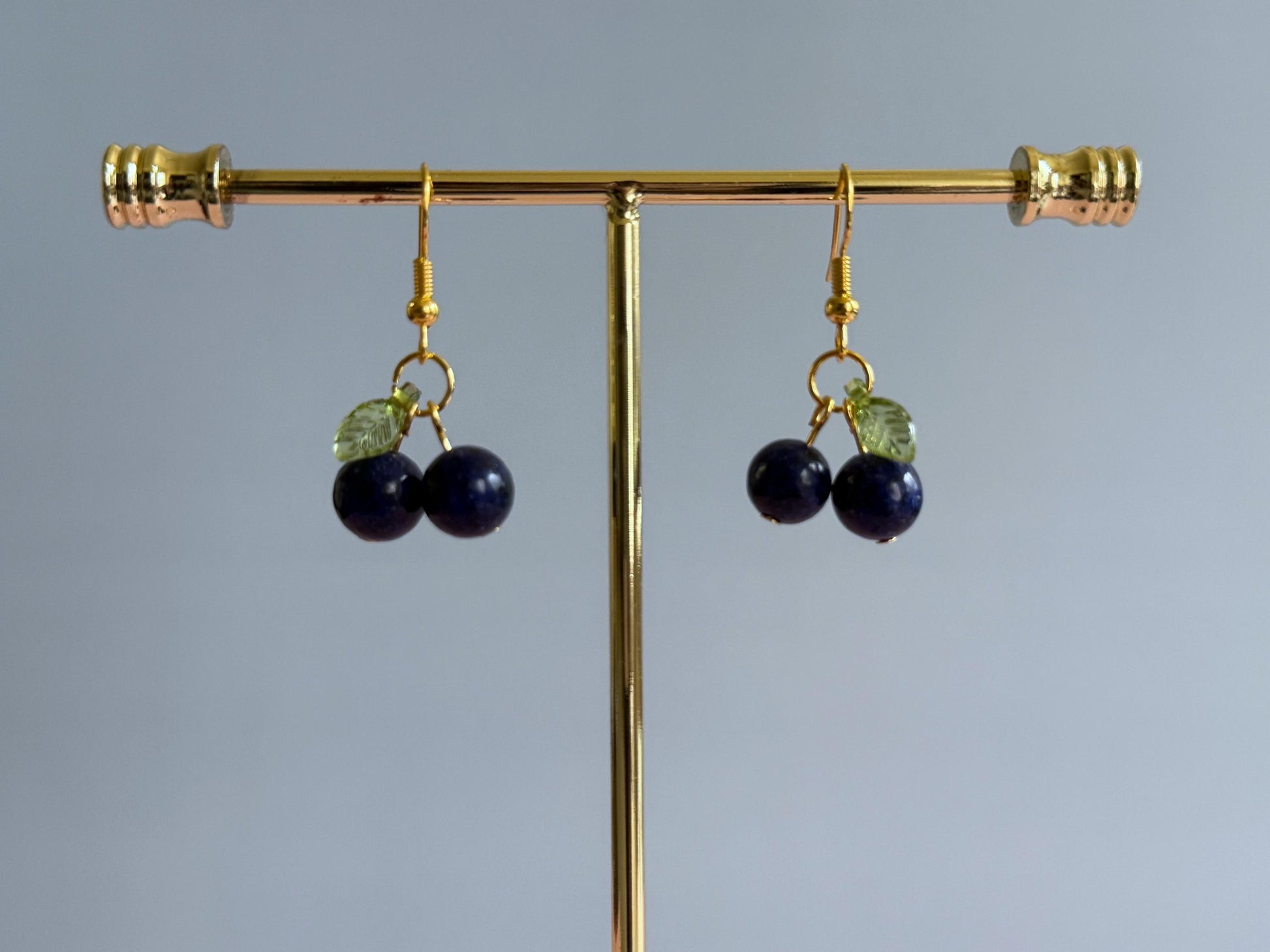 A pair of gold blueberry earrings on a stand. A pair of silver blueberry earrings hanging from a stand. The earrings feature a dark blue ball bead and an acrylic leaf bead.