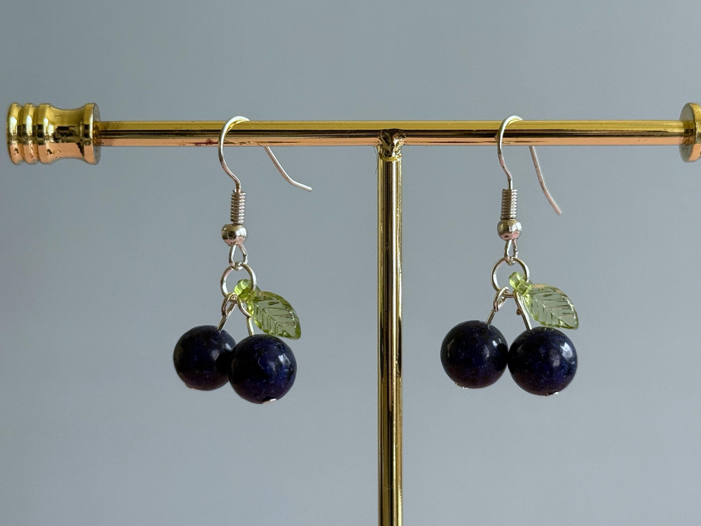 A pair of silver blueberry earrings on a stand. The earrings feature a dark blue ball bead and an acrylic leaf bead.