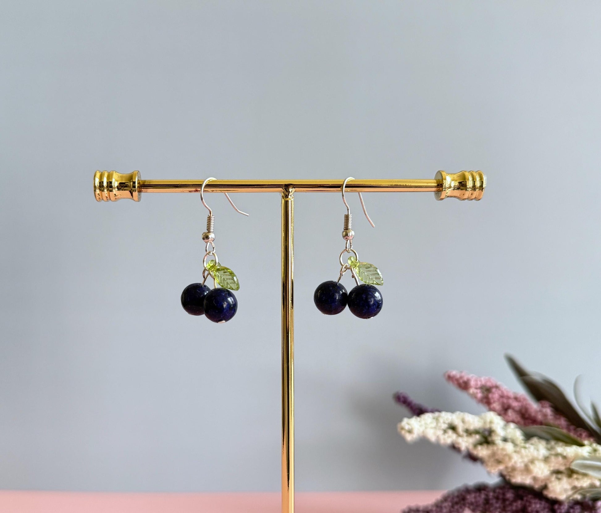 A pair of silver blueberry earrings on a stand. The earrings feature a dark blue ball bead and an acrylic leaf bead.
