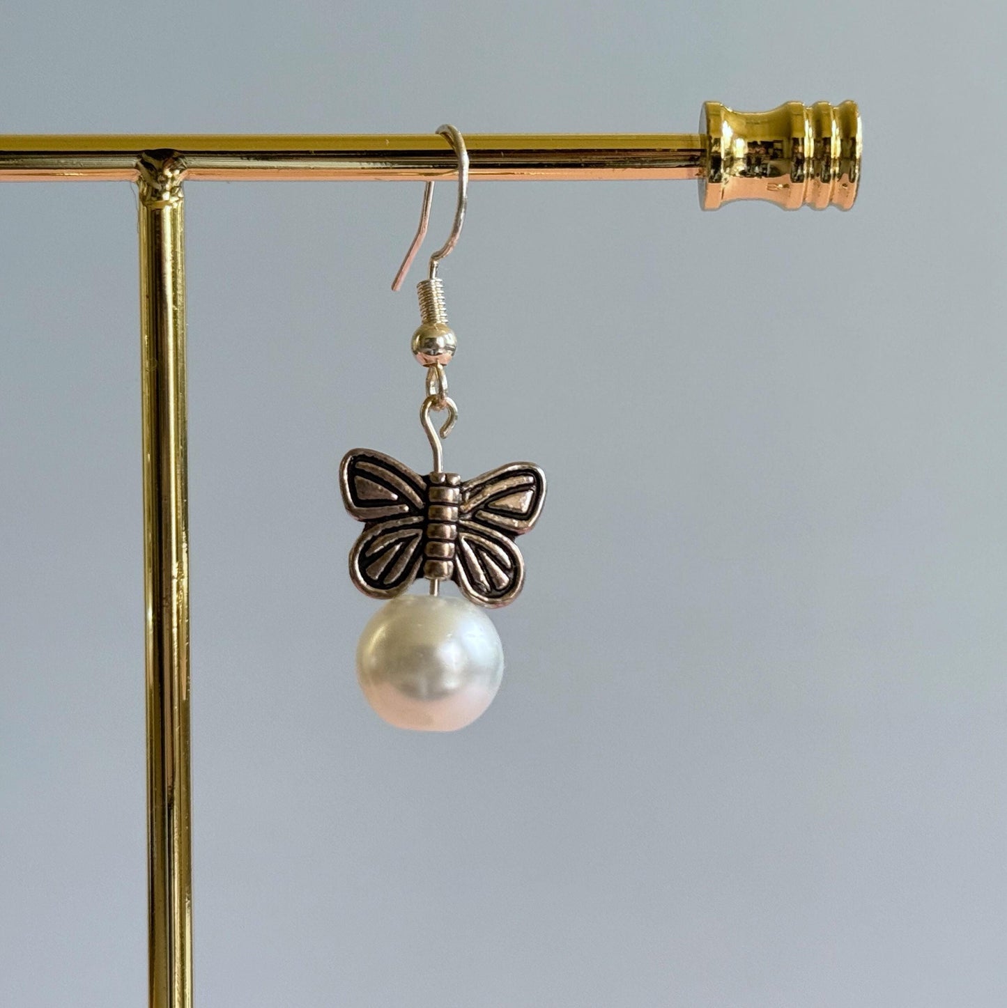 A close up of a silver butterfly earrings on a stand. The earrings feature metal butterflies and a pearl under the butterfly.