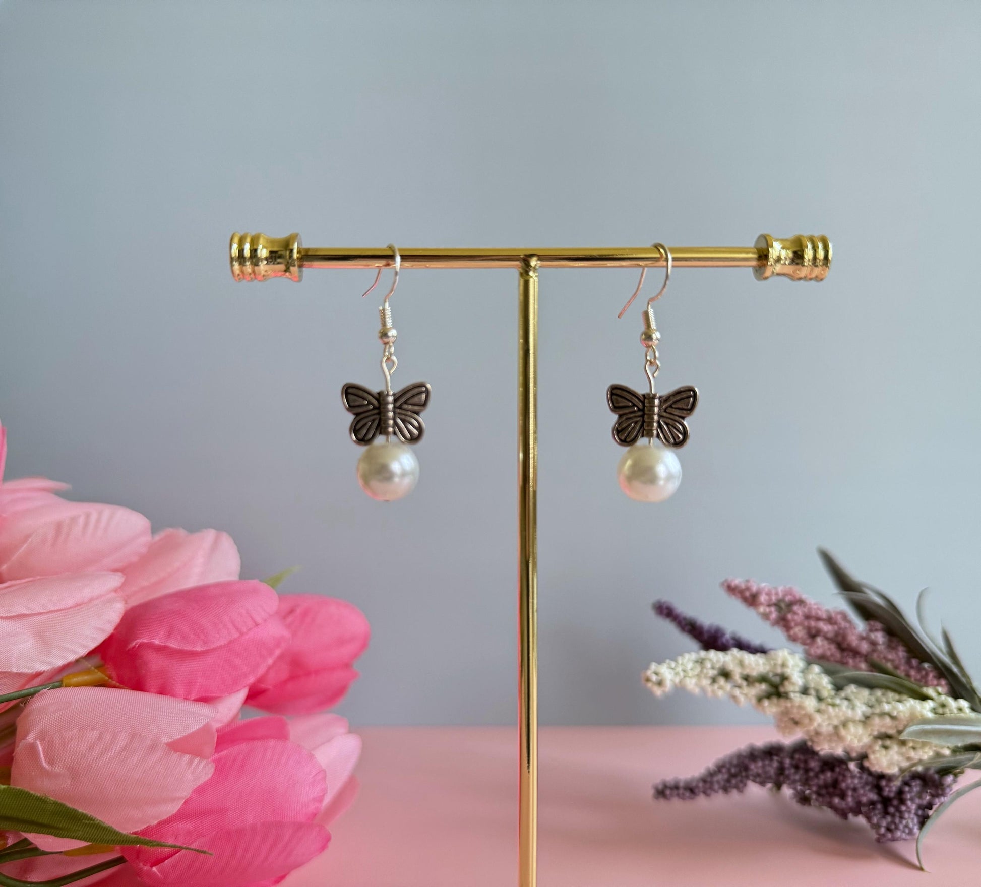 A pair of silver earrings butterfly earrings on a stand with flowers in the background. The earrings feature metal butterflies and a pearl under the butterfly.