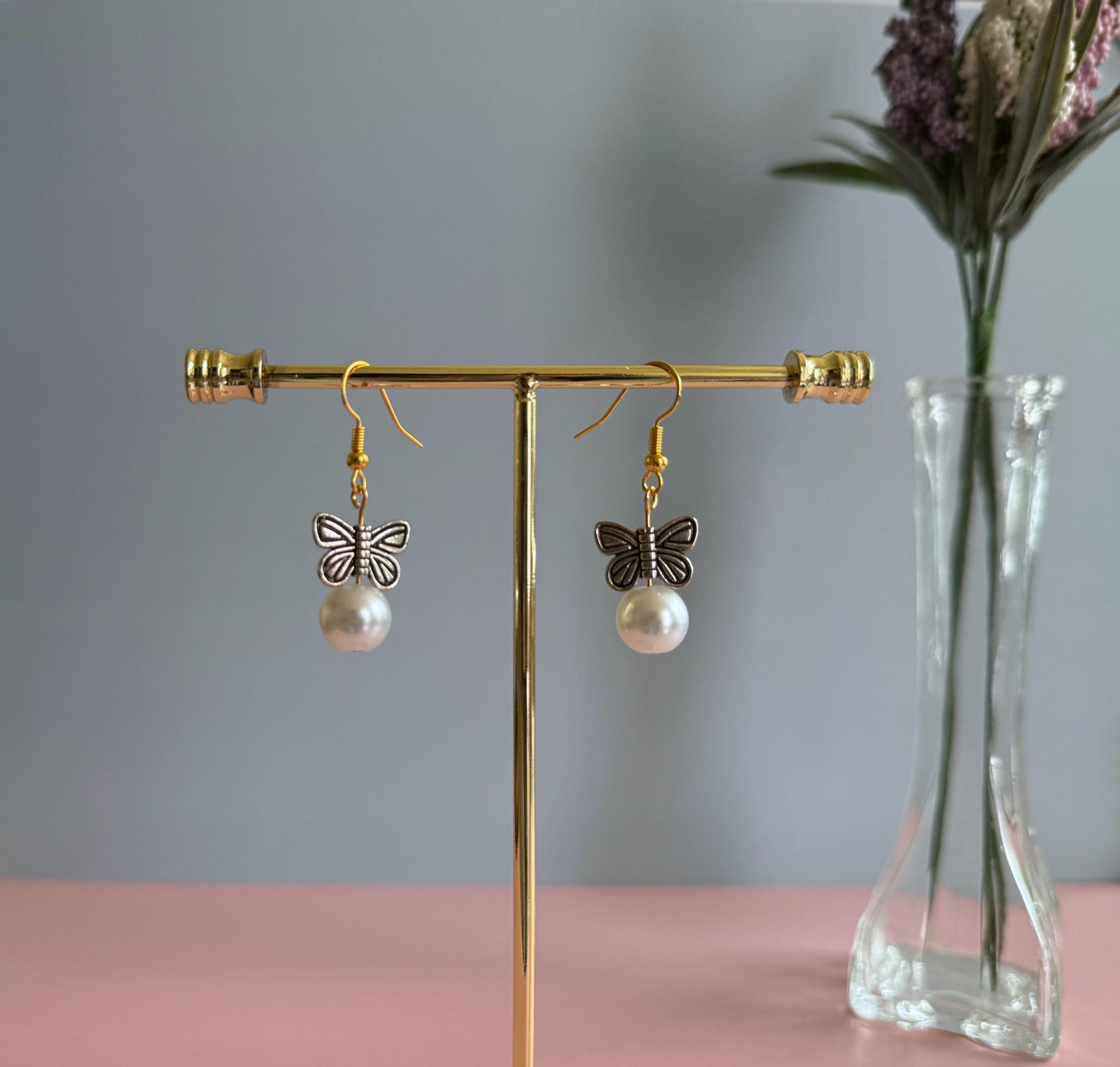 A pair of gold butterfly earrings hanging from a stand with purple flowers in the background. The earrings feature metal butterflies and a pearl under the butterfly.