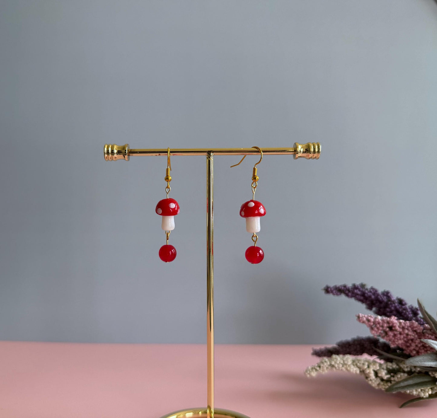 Red and white mushroom dangling earrings in gold with a red ball on the end on a stand with flowers in the background.