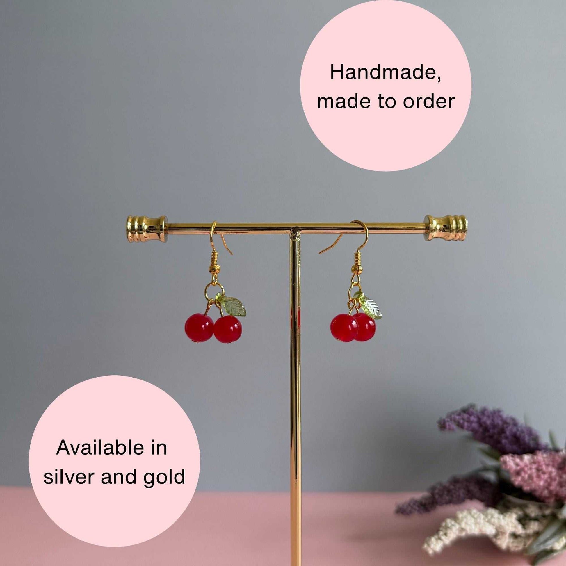 Red cherry earrings on a stand with text explaining that the earrings are in silver or gold and made to order